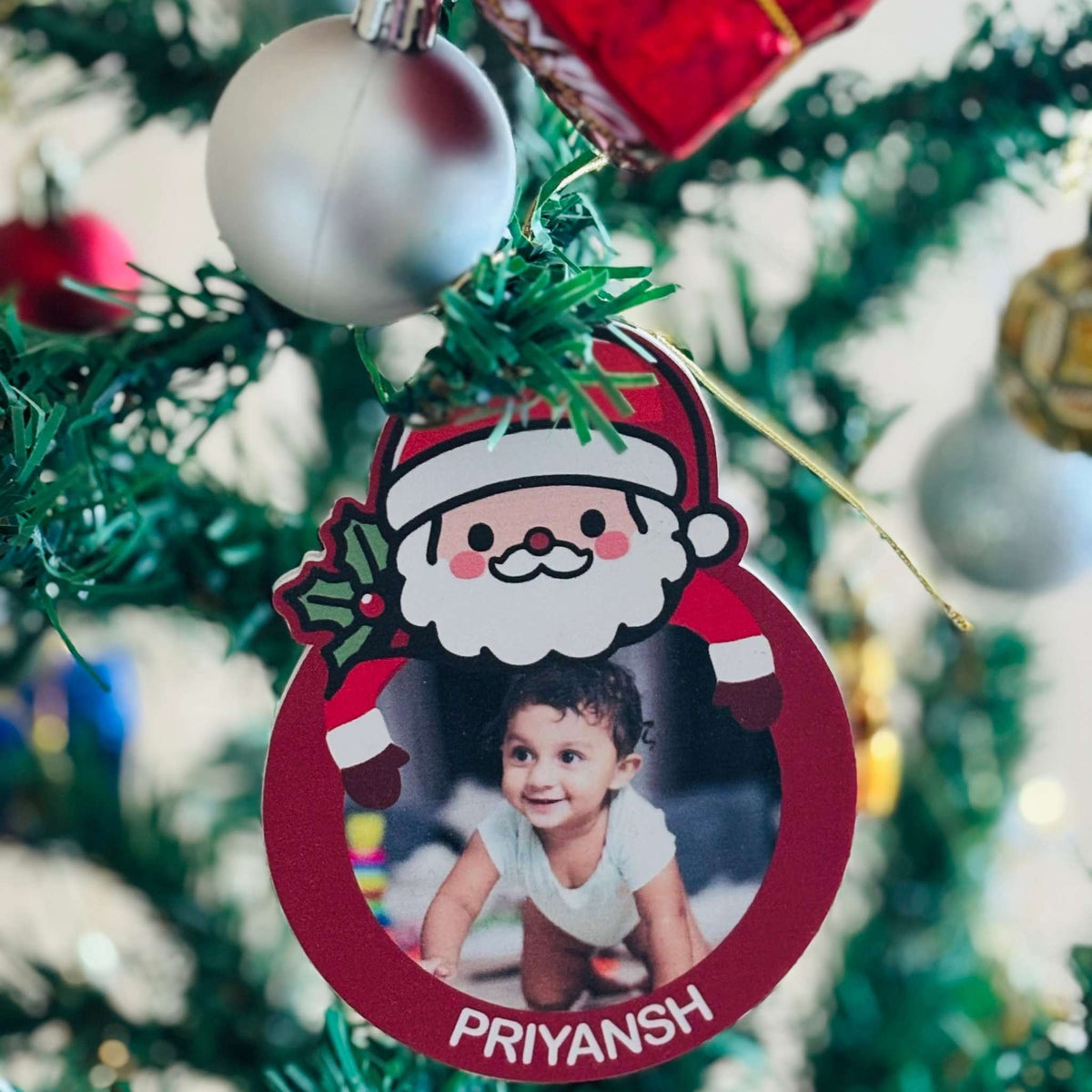 Personalized Christmas ornament with a child's photo and 'PRIYANSH' text, hanging on a decorated tree.