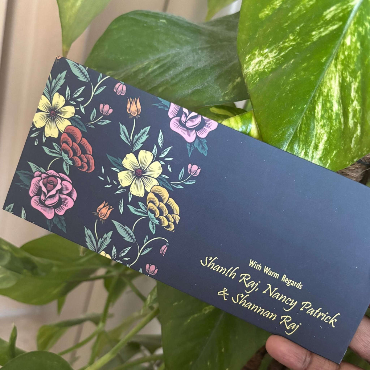 Floral design on a blue black envelope held by a hand with green leaves in the background
