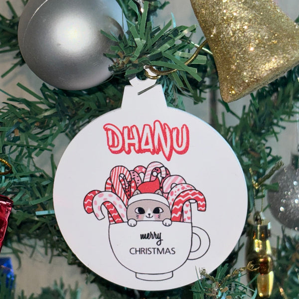 Christmas ornament with a cartoon cat holding candy canes and the word 'Dhanu' on a tree.