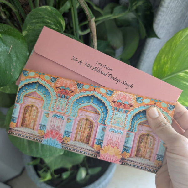 Decorative Jharokha design envelope with intricate designs held by a hand against a green plant background