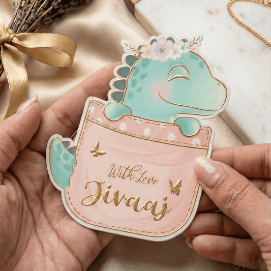 Hand holding a gift tag with a dino design and 'With love Jivaaj' text gold embossed placed on a soft fabric background.