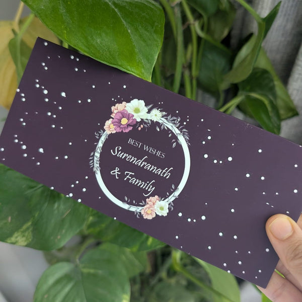 Hand holding a purple envelope with floral design and text in front of green leaves